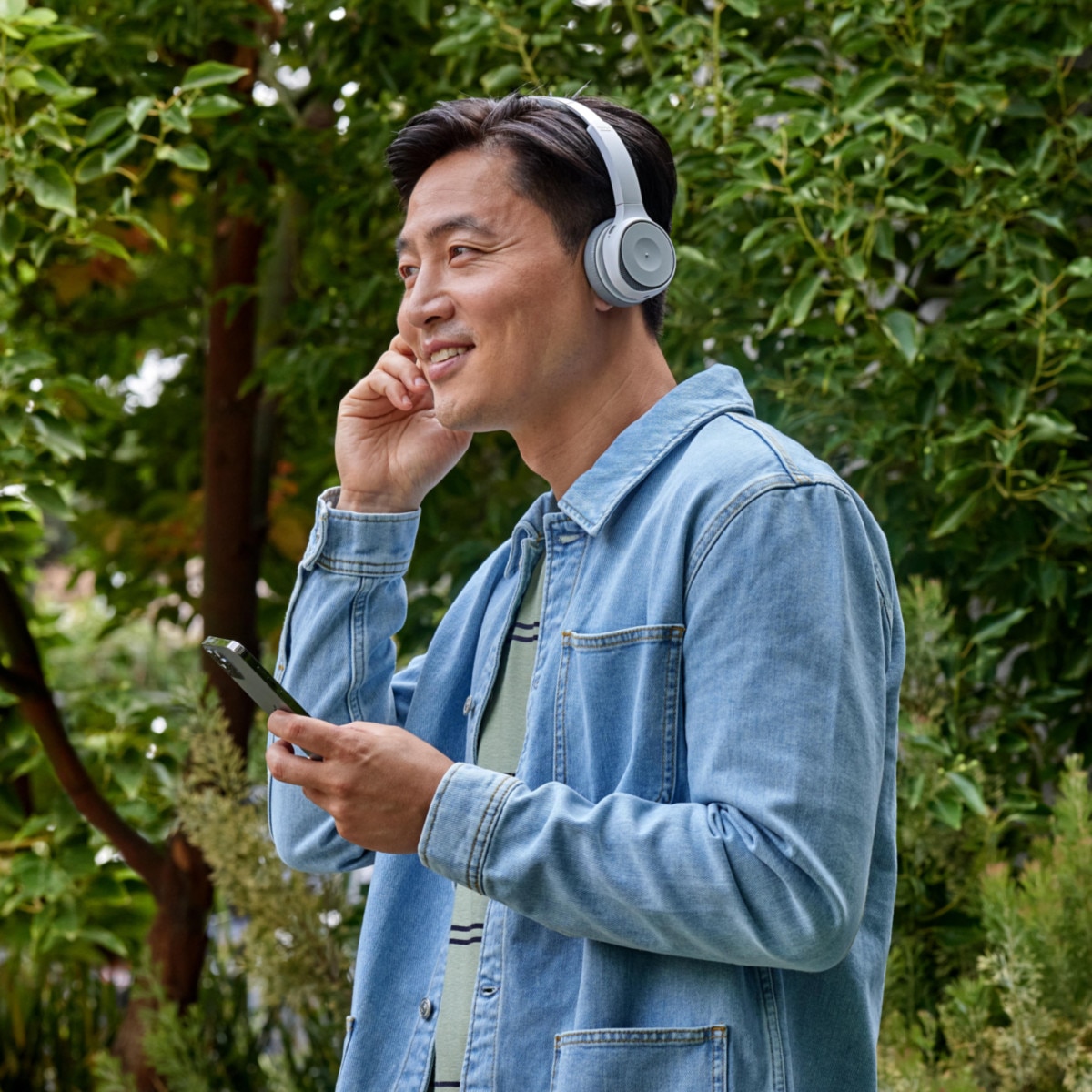 A person smiles while adjusting headphones and holding a mobile device outdoors.