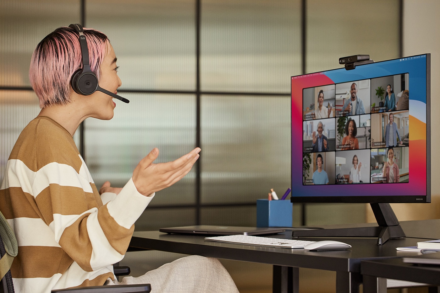 Woman wearing Cisco headset talks with team in grid view on video conference using Cisco desk camera.