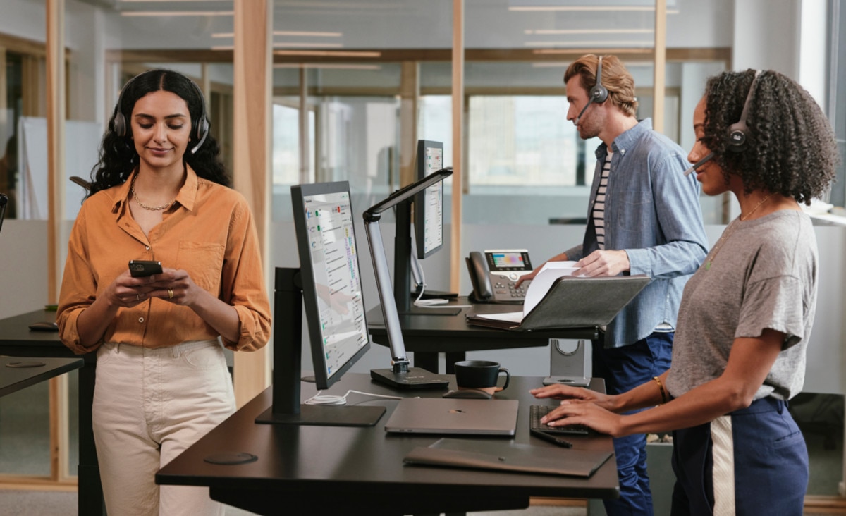 three people standing in an office, two are at standing desks. They all wear work headsets.