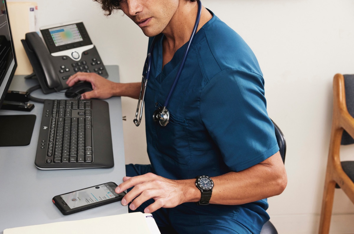 A medical professional uses VoIP while sitting at a desk that includes an IP phone, a smartphone, and a monitor.