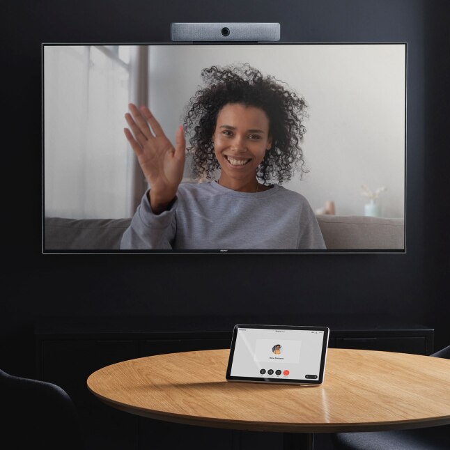 A person smiles and waves from a wall-mounted monitor.