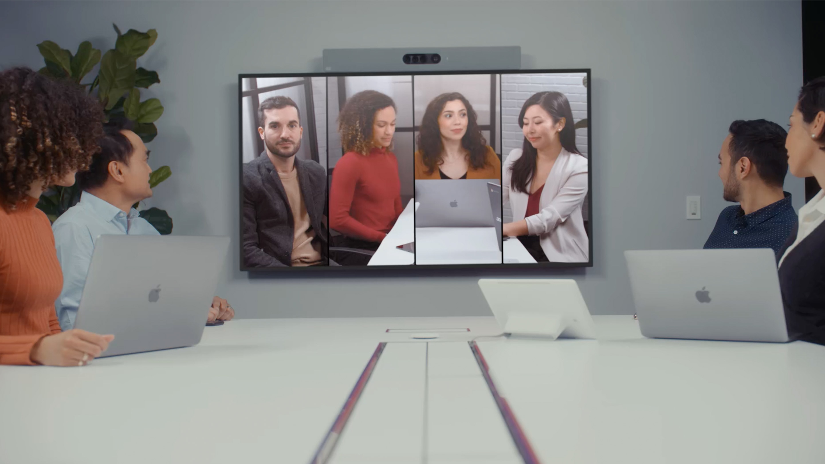 A large display with the Quad Camera Bar mounted to the top shows employees in Frames view during a video conference, while in-person team looks on in a conference room.