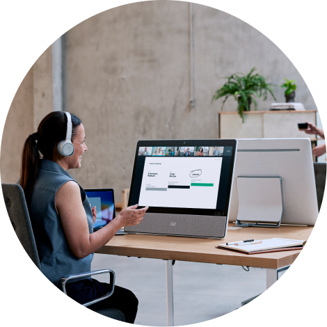 Sitting at a desk in an open office, a professional wearing a headset video conferences with several colleagues.