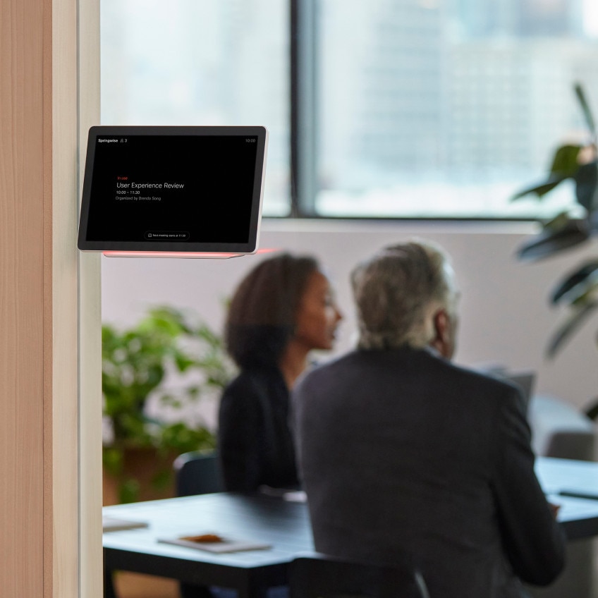 2 people collaborate in a conference room. A Webex Room Navigator outside the room shows the room is in use until 11:30.