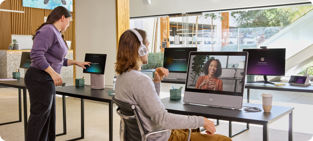 Sitting at a desk in an open office, a professional wearing a headset video conferences with a colleague.