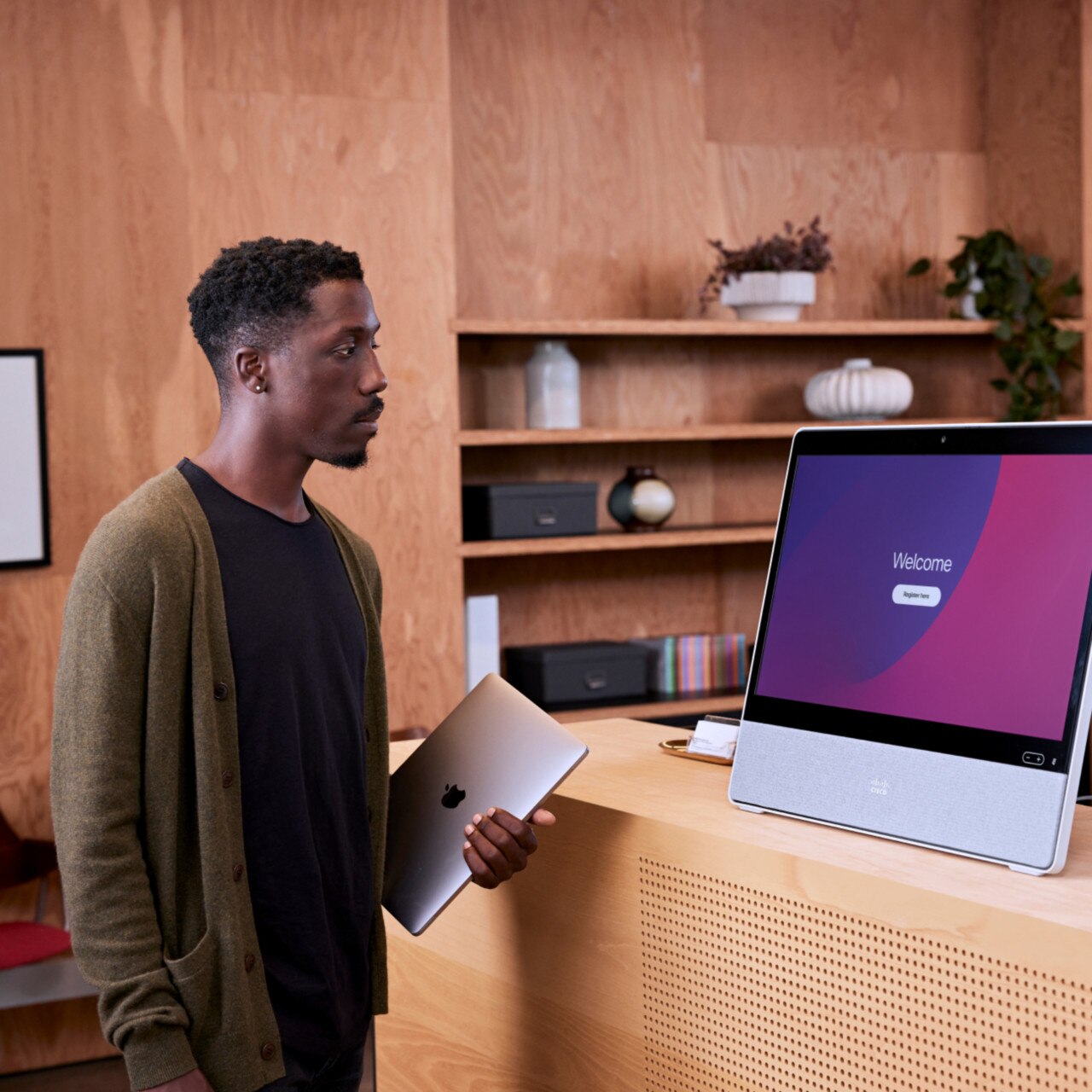 Person pauses at the reception desk in the office, looking at a screen that says Welcome and Register here.
