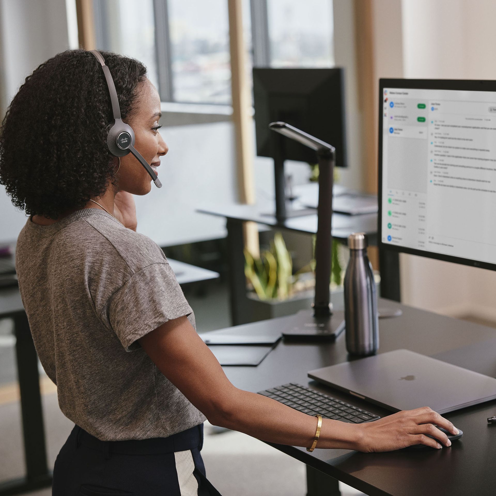 A smiling person in a headset works at a computer at a standing desk in an open office.