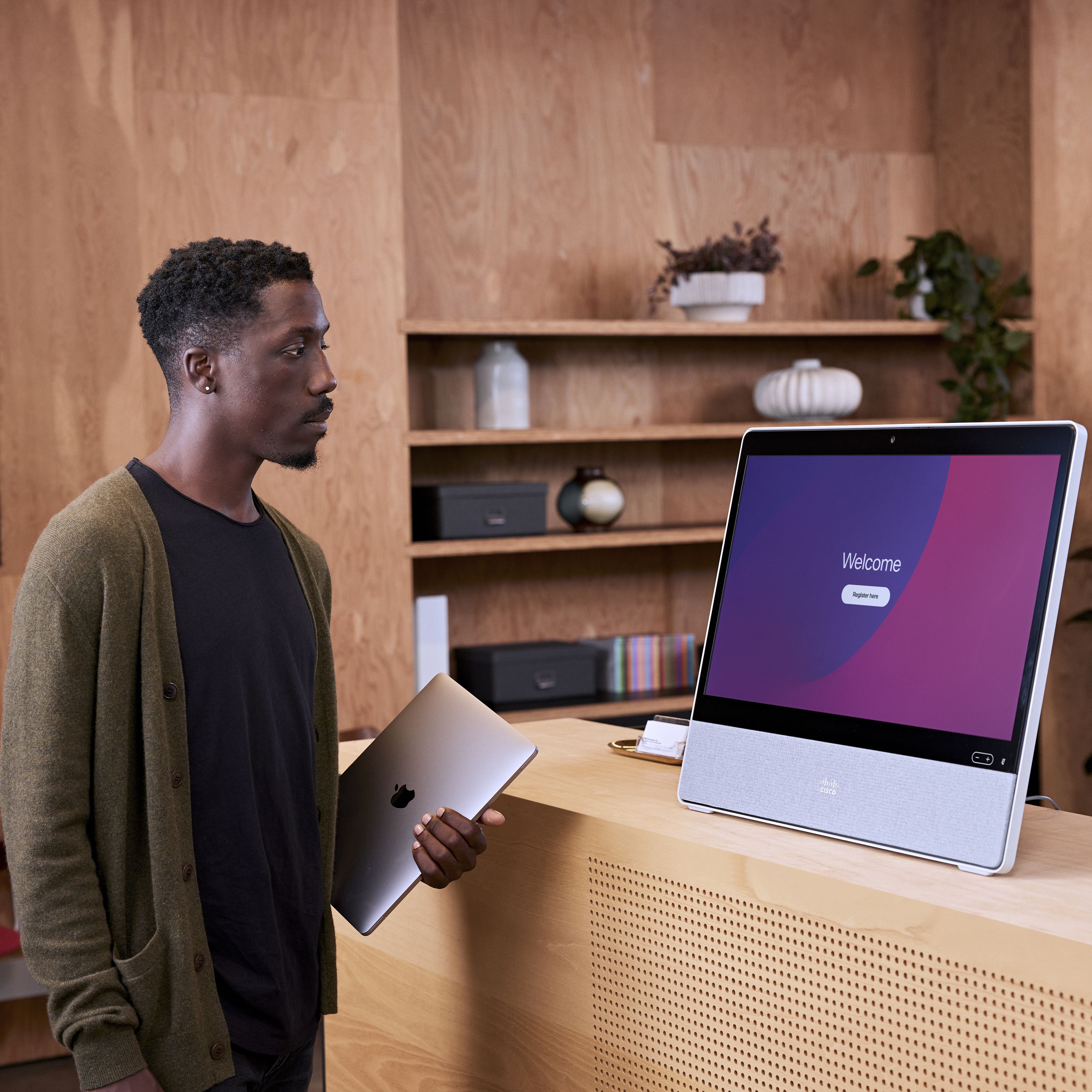 A person holding a laptop pauses at a Cisco Desk on a counter in the office lobby. Screen displays a welcome message.