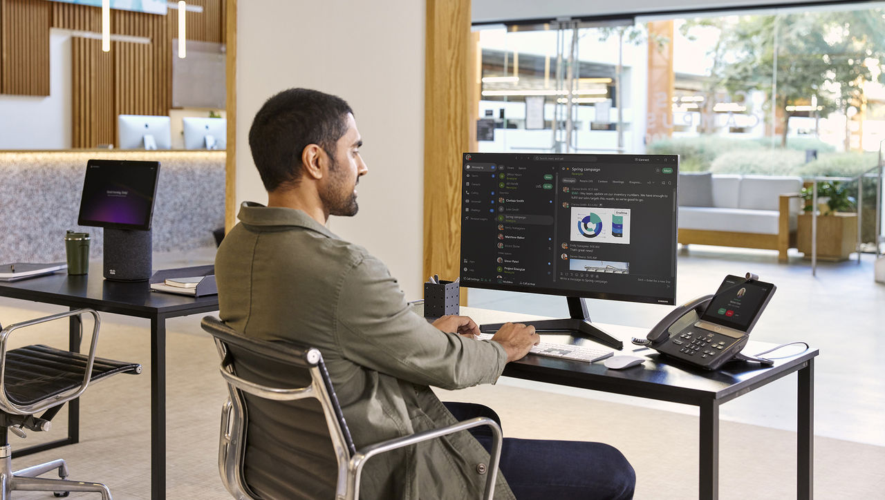 A man sitting at a desk in a bright office, working on a computer displaying a messaging app with a pie chart, with a Cisco desk phone nearby.