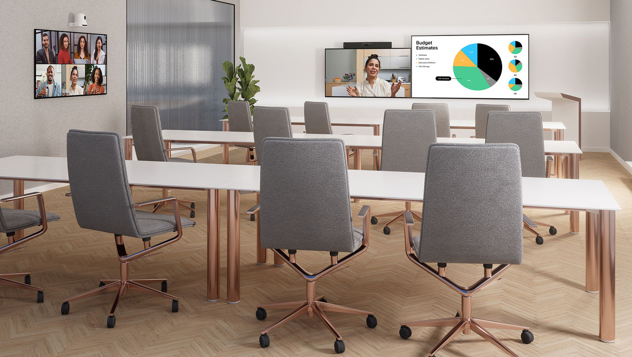 A modern, well-lit conference room with grey chairs, a long white table, and multiple wall-mounted screens displaying a video call and a "Budget Estimates" pie chart.