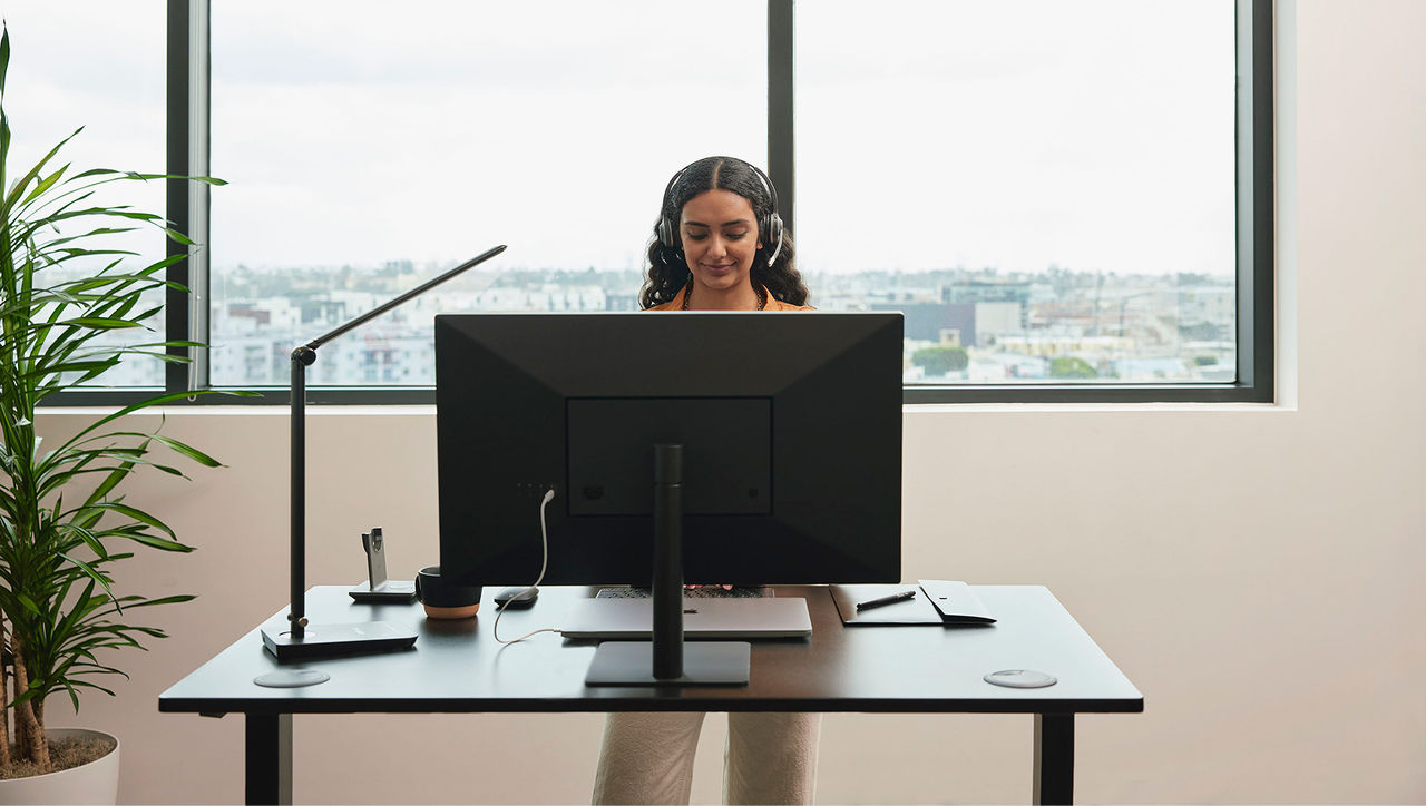 A woman wearing a headset, standing at a desk and working on a computer monitor in front of a window with a city view.