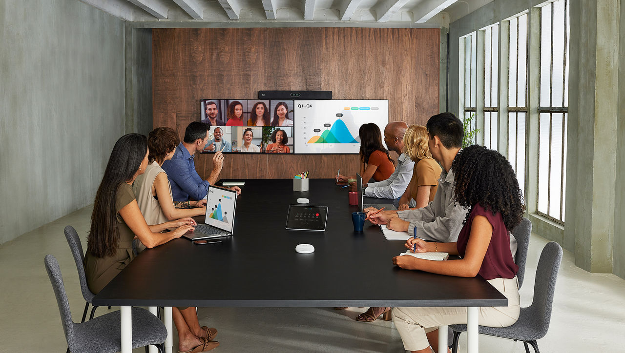 A team in a conference room participating in a hybrid meeting, with a large screen displaying business analytics.