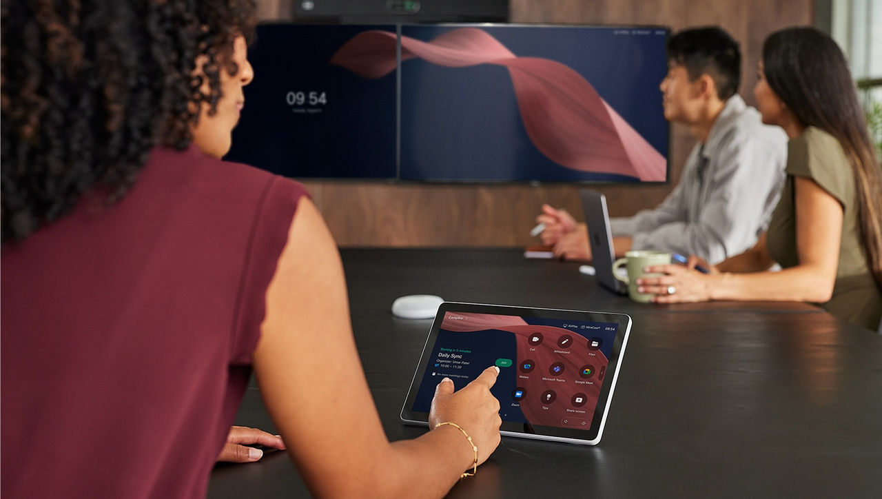 A person in a meeting room interacting with a tabletop touch-screen device, with colleagues seated in the background.