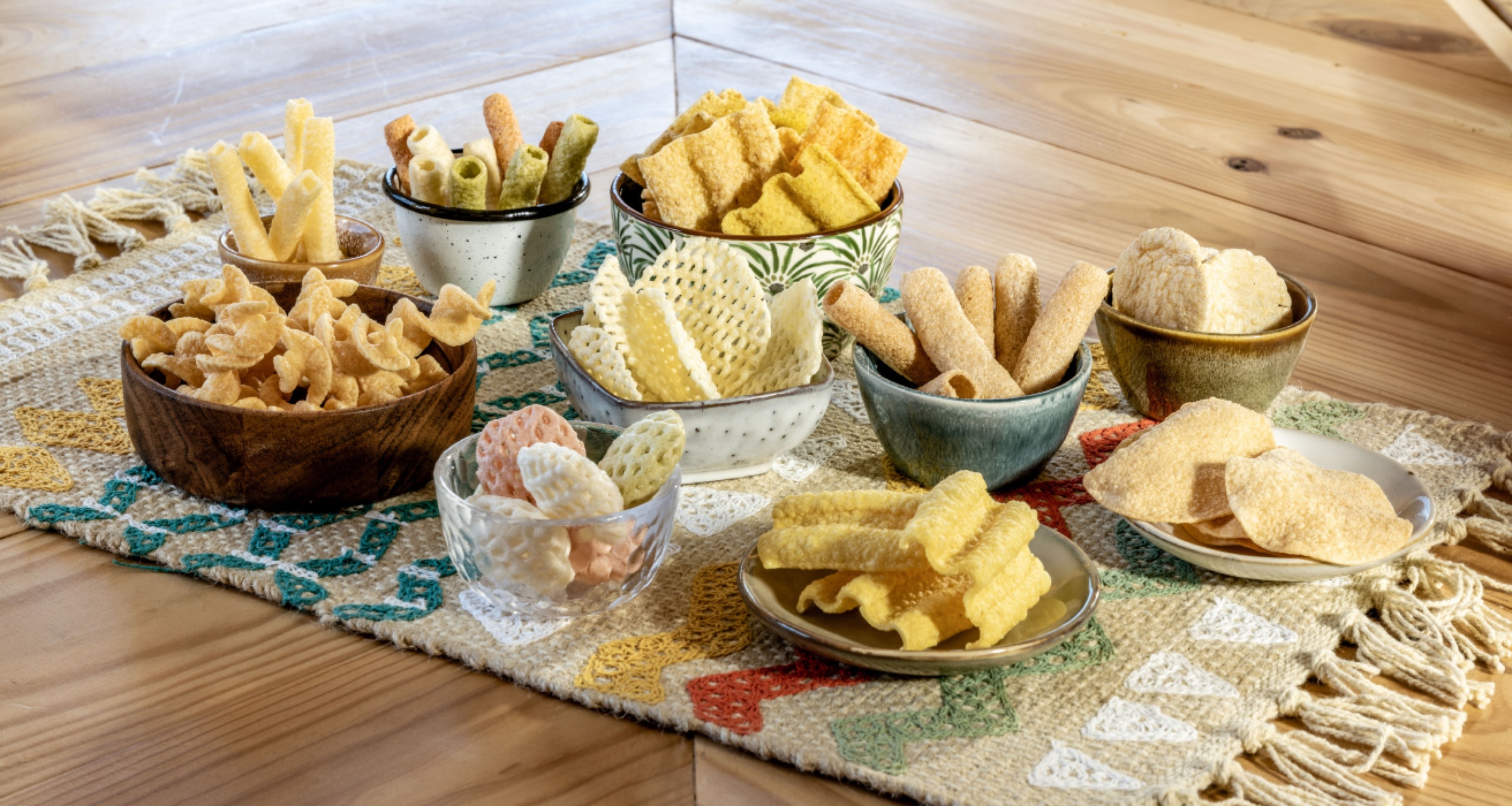 Several types of chips and snacks on little plates and bowls are displayed on colorful woven placemat atop a wooden table.
