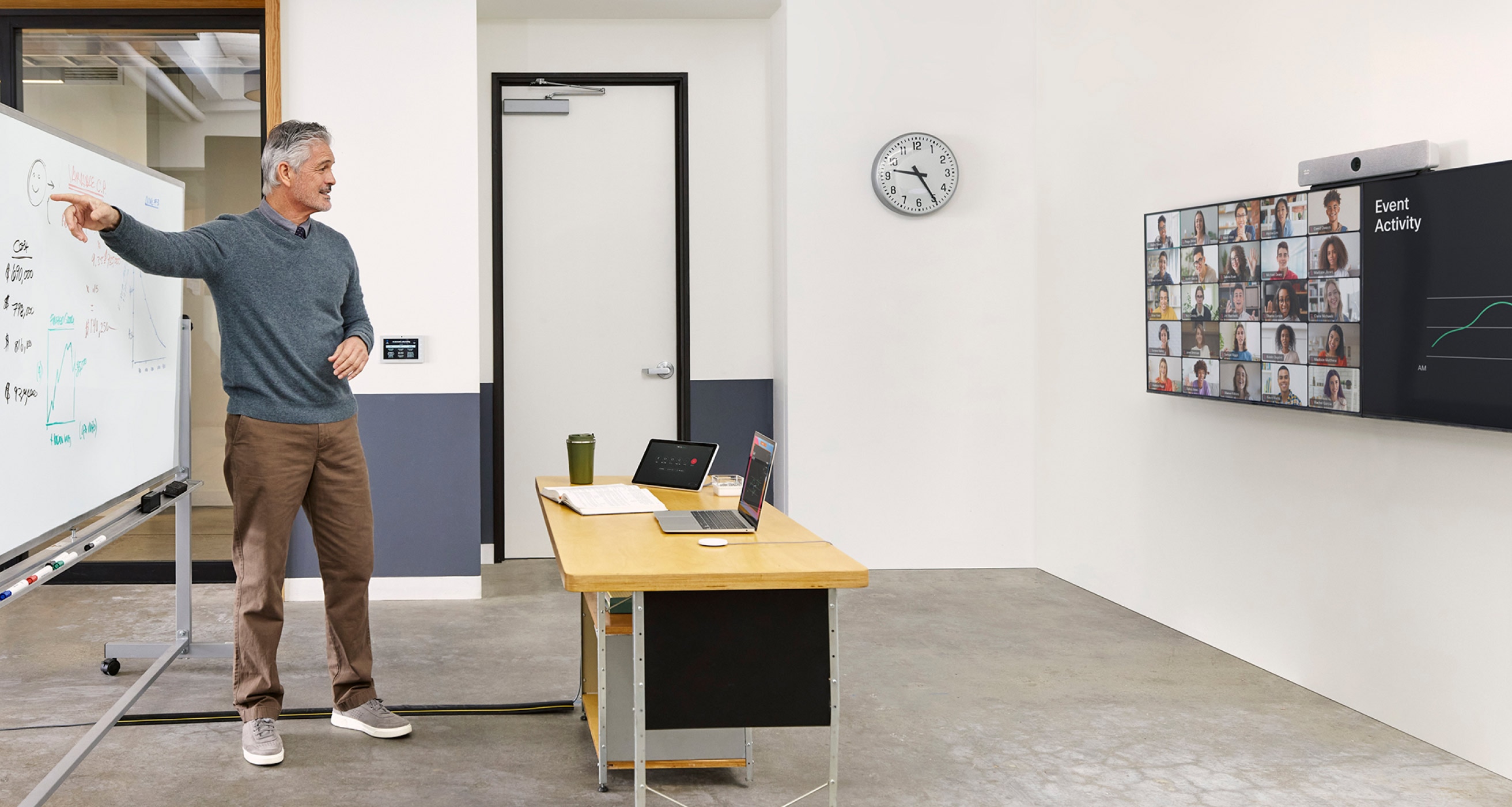 A teacher stands at a whiteboard in a classroom, teaching a class of remote students who can be seen on a screen in the room.