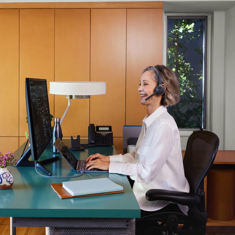 A professional wearing a headset smiles while typing on a laptop.